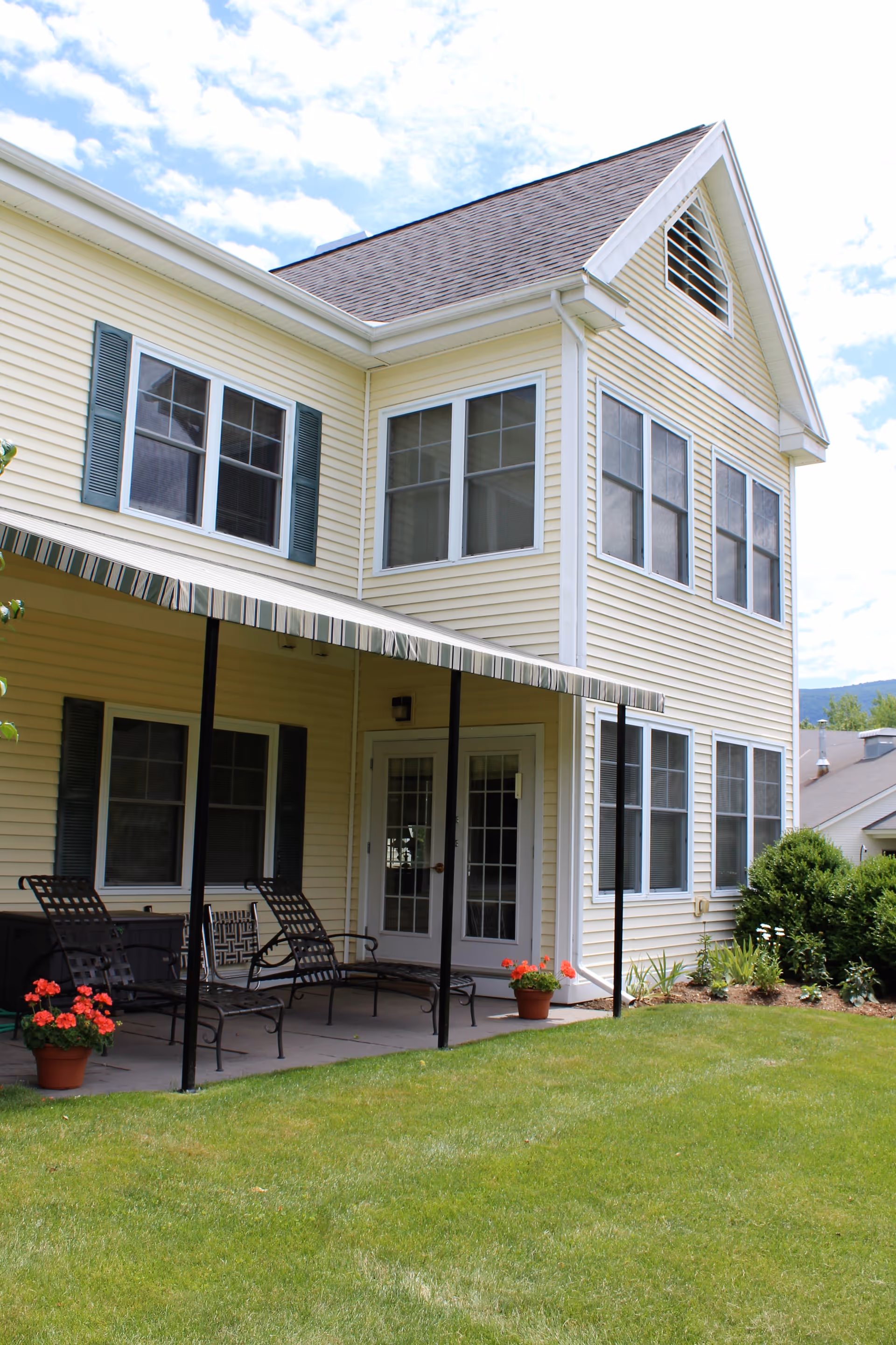 Outdoor patio area of a yellow two-story building with multiple windows and a striped awning. The patio has metal chairs and potted red flowers, with a well-maintained green lawn in the foreground.