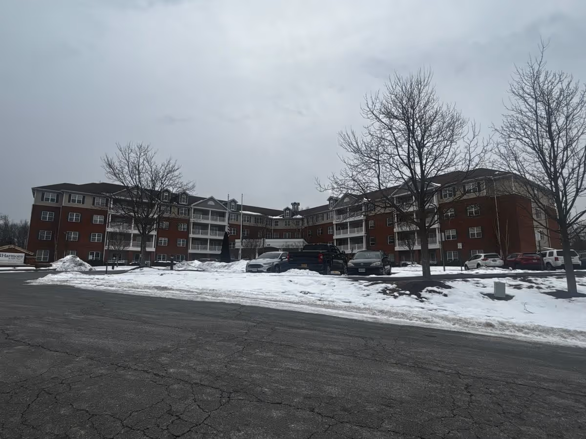Brick multi-story senior living building with a snow-covered parking lot and leafless trees under a cloudy sky.