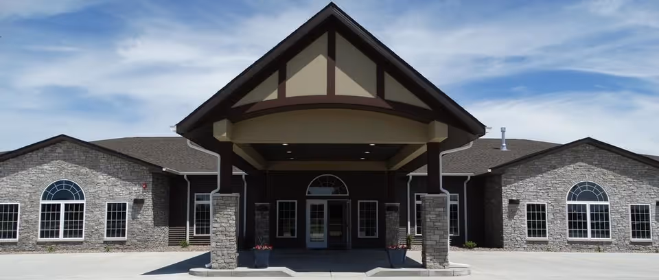 Front entrance of a single-story stone-faced assisted living building with a covered porte-cochère and arched windows.