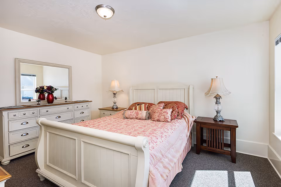A bright bedroom with a white wooden bed frame and a pink patterned bedspread. The bed is adorned with multiple decorative pillows. On either side of the bed are two bedside tables, each with a decorative lamp. To the left is a white dresser with multiple drawers and a large mirror above it, with a vase of flowers on top. The room has white walls and a window letting in natural light.