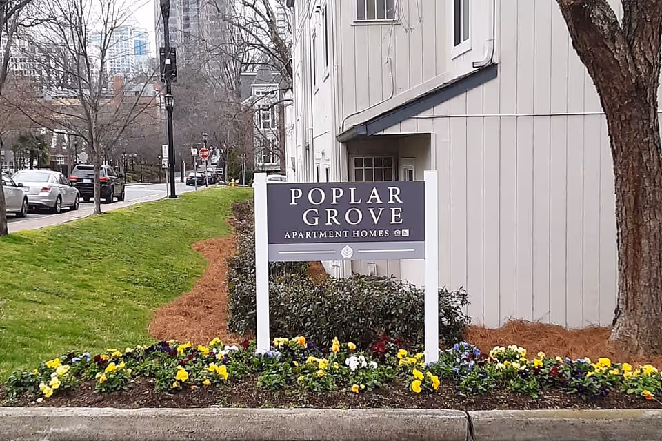 A sign for Poplar Grove Apartment Homes is displayed in front of a beige building with a tree and flower bed containing yellow, purple, and white flowers. A street with parked cars and leafless trees is visible in the background.