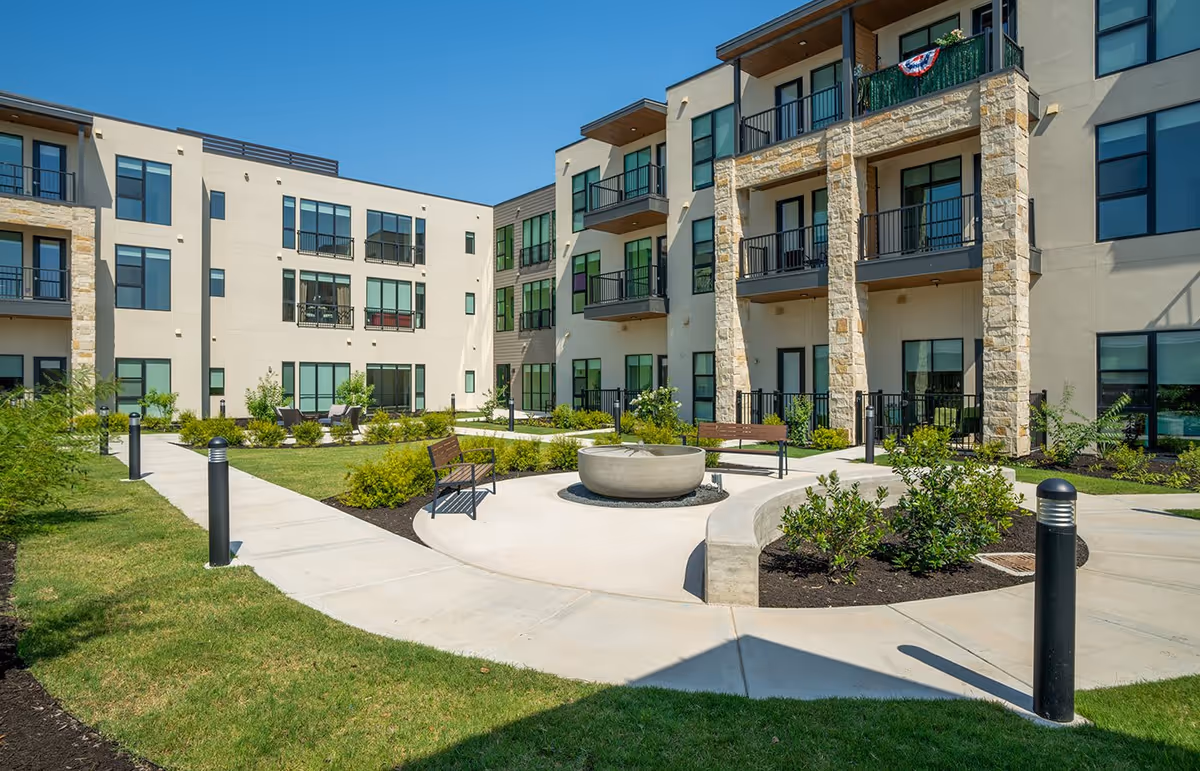 Outdoor courtyard area of The Hacienda at Georgetown featuring a circular concrete seating area with a central fire pit, surrounded by benches and landscaped greenery. The courtyard is enclosed by a modern three-story building with large windows and balconies, under a clear blue sky.