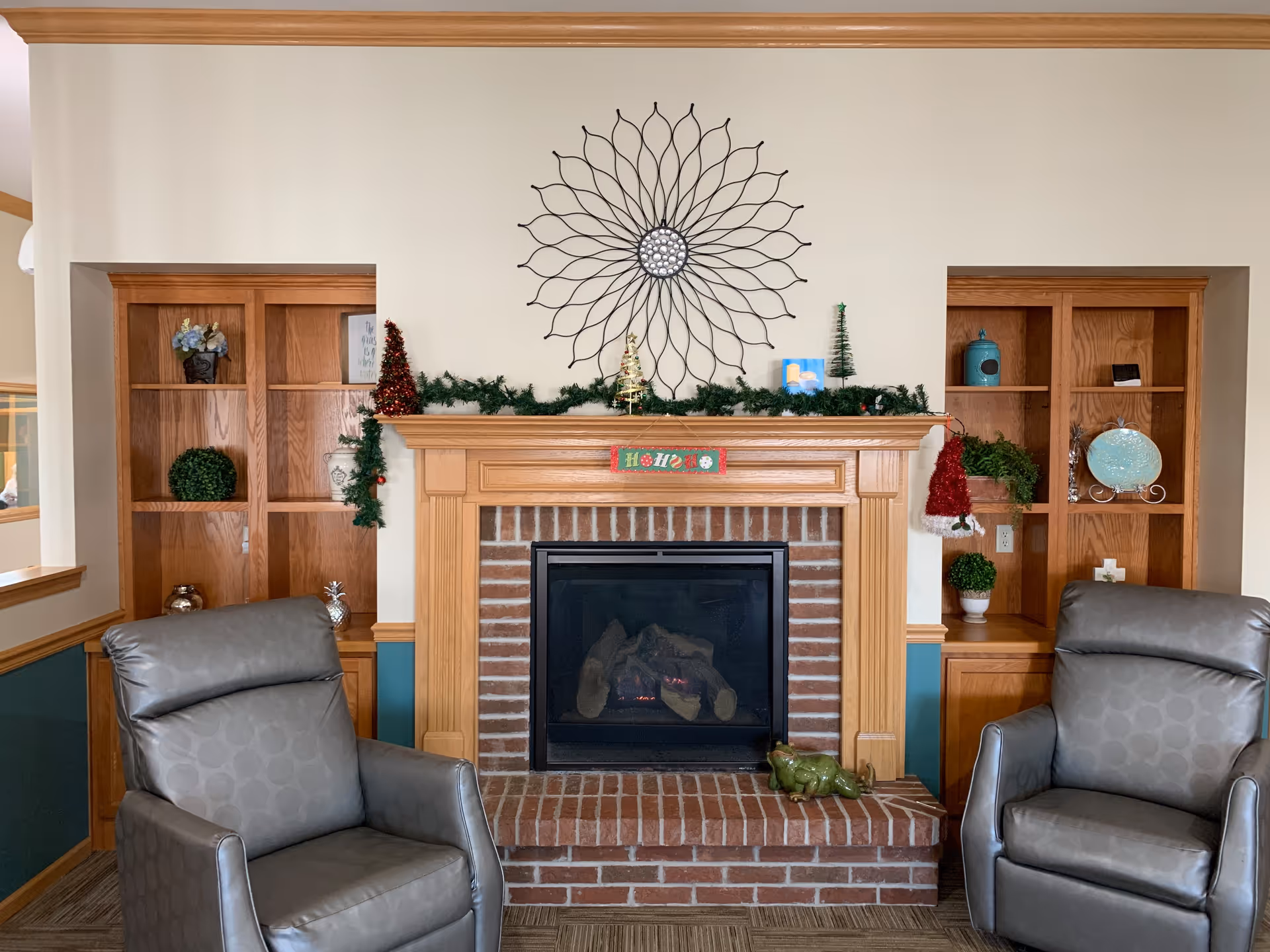 A cozy living room area featuring a brick fireplace with a wooden mantel decorated with Christmas garland, small Christmas trees, and a HO HO HO sign. Above the mantel is a large decorative metal wall art piece. On either side of the fireplace are built-in wooden shelves with various decorative items including plants, vases, and plates. Two gray leather armchairs are positioned in front of the fireplace.