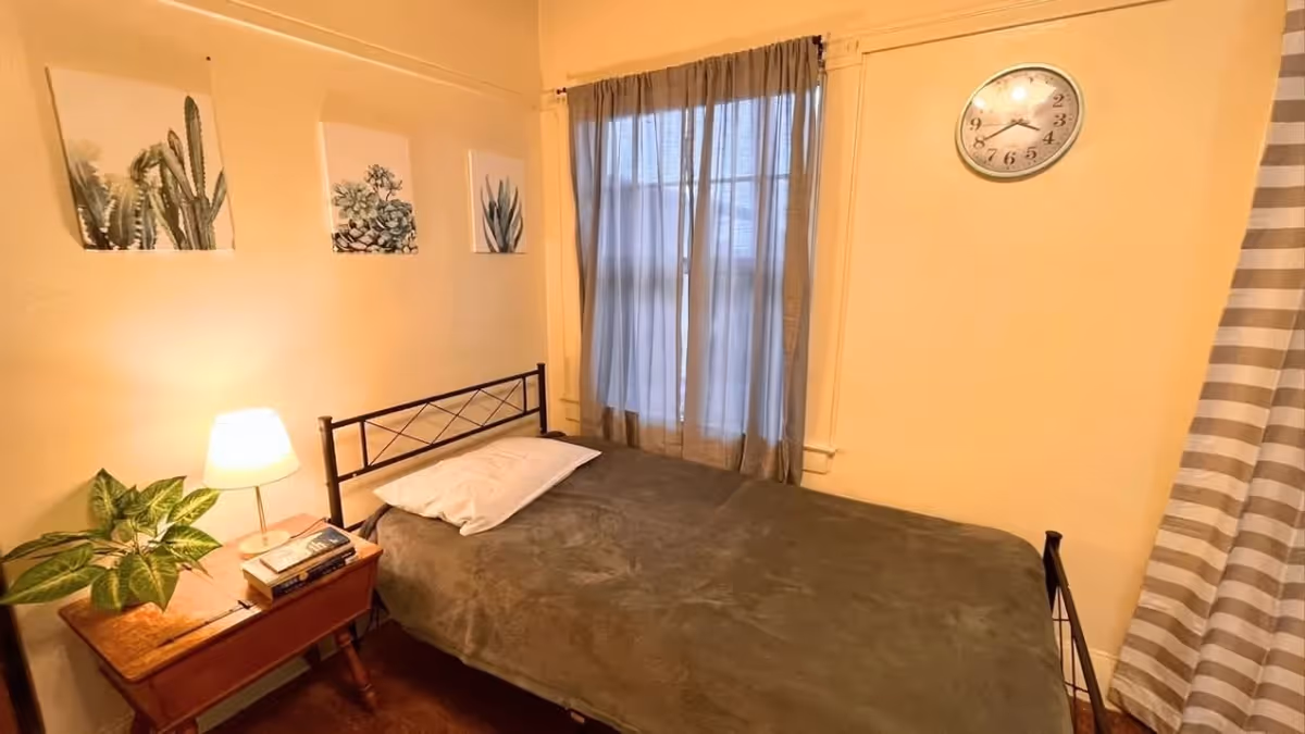 A small bedroom with a single bed covered in a dark blanket and a white pillow. Next to the bed is a wooden nightstand with a lamp, a plant, and some books. Three framed botanical prints hang on the wall above the bed. A window with sheer curtains is behind the bed, and a round wall clock is mounted on the adjacent wall.