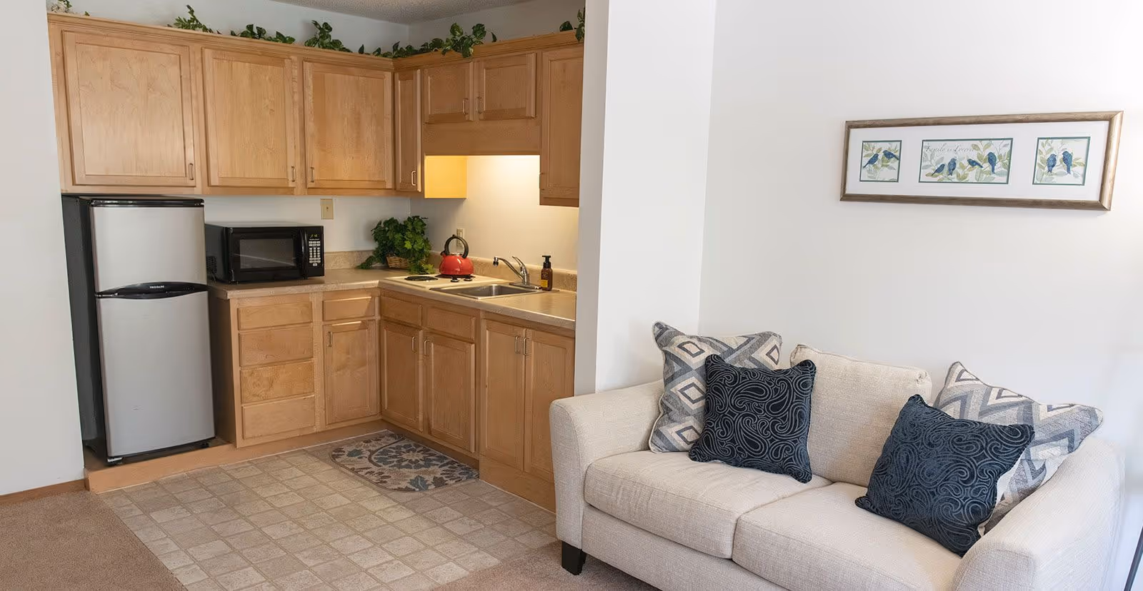 A small kitchen area with light wood cabinets, a stainless steel refrigerator, a microwave, a red kettle on the stove, and a sink. Adjacent to the kitchen is a beige couch with patterned throw pillows and a framed artwork of birds on the wall above it.