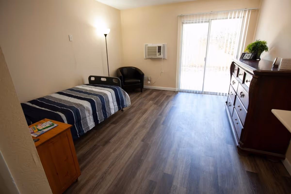 Furnished senior bedroom with a single bed, wooden dresser, black armchair, floor lamp, window air conditioner and sliding glass door.