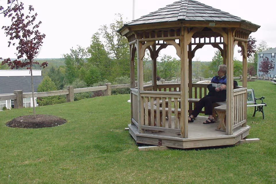 An elderly person sitting inside a wooden gazebo on a grassy lawn with trees and buildings in the background.