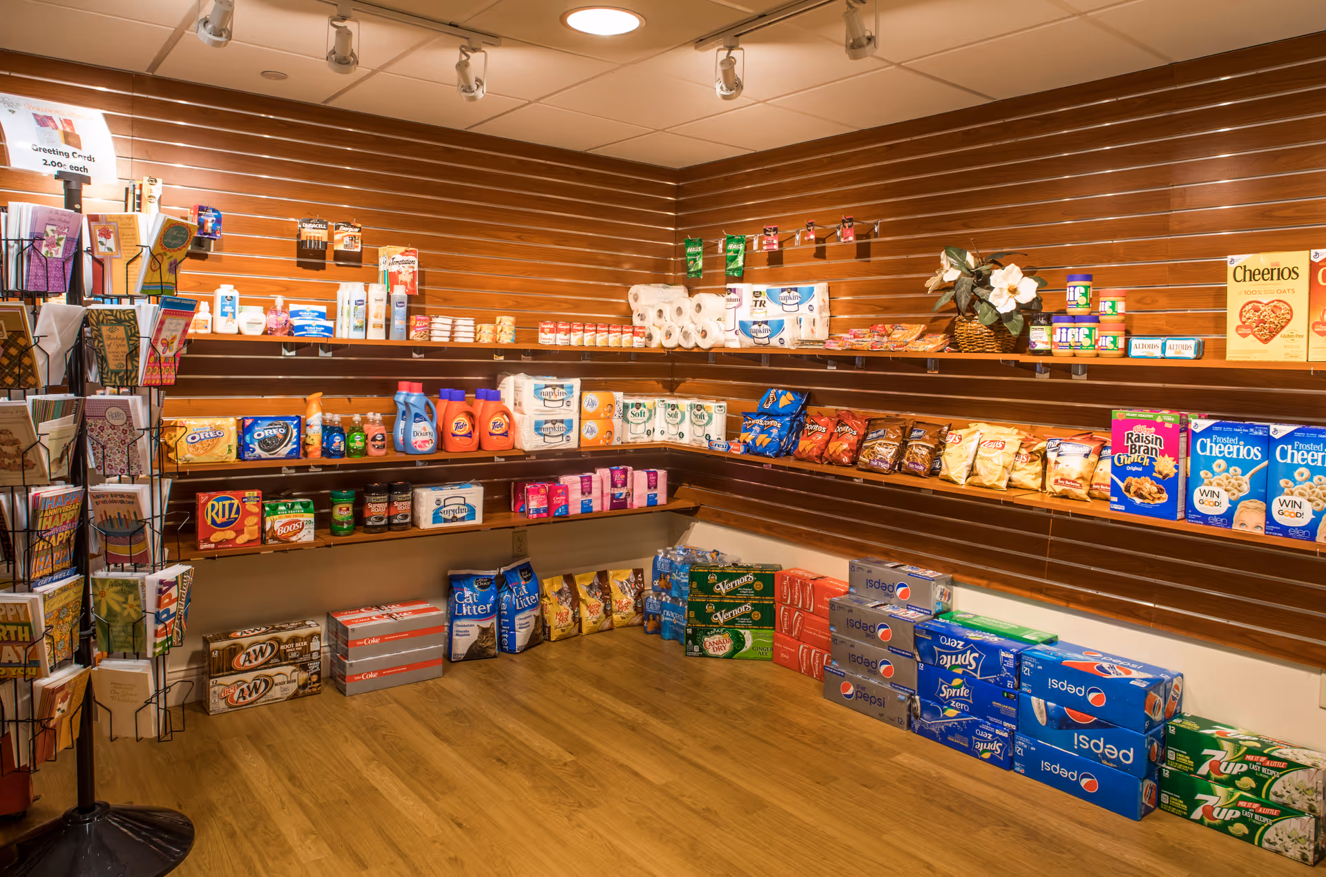 Small convenience store corner inside a facility with wooden shelves stocked with various grocery items including snacks, cereals, canned goods, cleaning products, and beverages. A rotating rack with greeting cards is visible on the left side. The floor is wooden and the lighting is warm with ceiling spotlights.