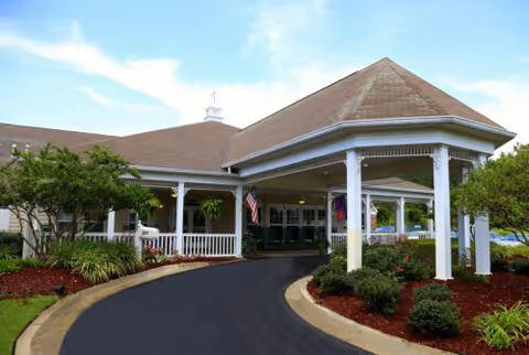 Exterior view of a single-story building with a covered entrance supported by white columns. The building has a brown roof and beige walls. There are landscaped bushes and trees along the curved driveway leading to the entrance. An American flag is displayed near the entrance under the covered area.