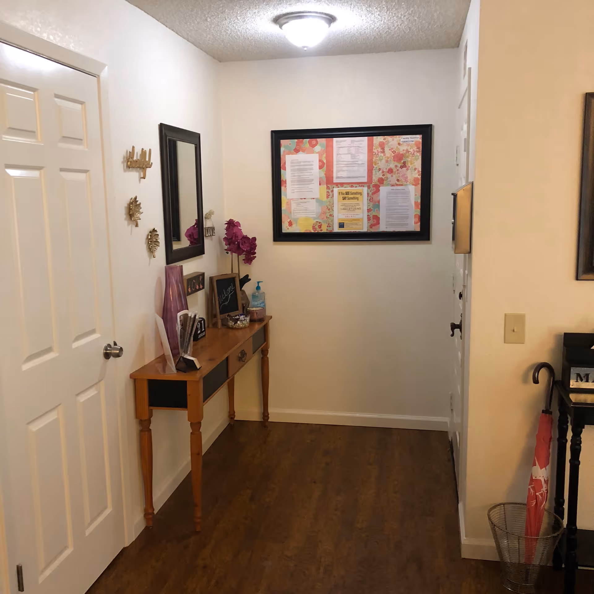 A hallway with wooden flooring and cream-colored walls. On the left side, there is a wooden console table with decorative items including a purple vase, a small chalkboard, and a bottle of hand sanitizer. Above the table is a rectangular mirror and some wall decorations. At the end of the hallway, a bulletin board with various notices is mounted on the wall. On the right side, there is a black stand with an umbrella holder containing a pink umbrella.