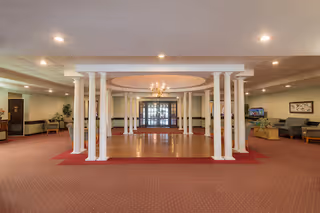 Spacious interior common area with a red carpet and a central section featuring white columns and a chandelier. Seating areas with sofas and chairs are visible on the right side, along with a television. The far end has double glass doors and some framed artwork on the walls.