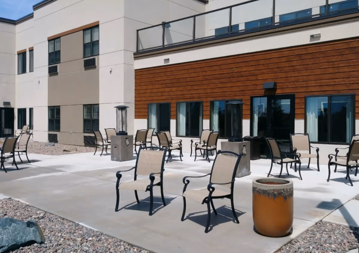 Outdoor patio area at Cambridge Senior Living- Rice Lake with multiple beige and black metal chairs arranged on a concrete surface, surrounded by a building with beige and brown exterior walls and several windows.