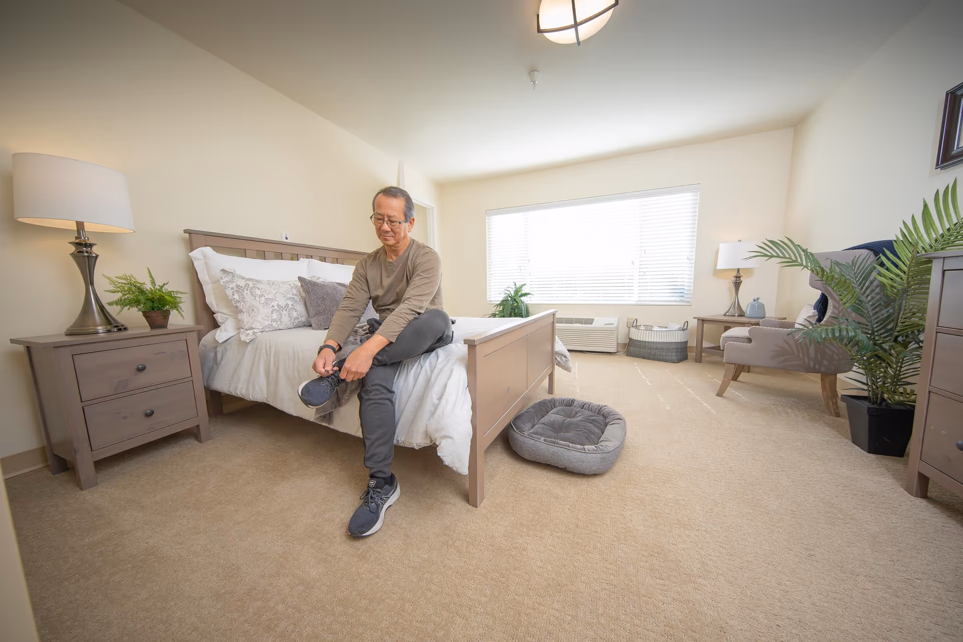 An older man sits on the edge of a bright, furnished bedroom bed tying his shoe.