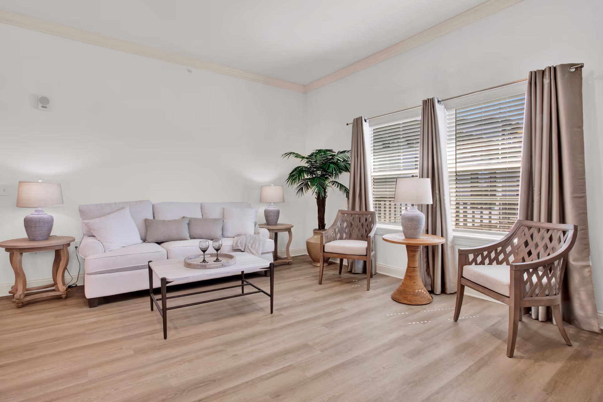 A bright living room with a white sofa adorned with cushions, two wooden side tables each with a lamp, two wooden armchairs with cushions, a wooden coffee table with three wine glasses on a tray, large windows with beige curtains, and a potted plant in the corner.