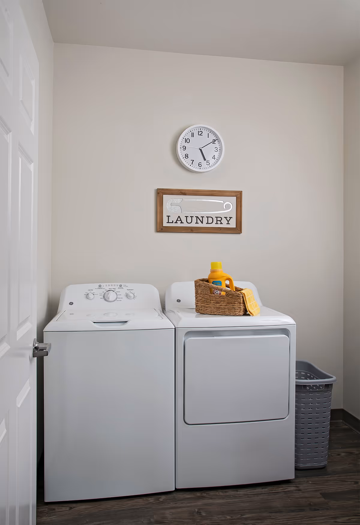 A laundry room with a white washing machine and dryer side by side. On top of the dryer is a wicker basket containing a yellow bottle of detergent and a yellow cloth. Above the machines, a wall clock shows the time as 5:10, and a framed sign with a safety pin illustration reads 'LAUNDRY'. A gray laundry basket is placed on the floor to the right of the dryer. The room has light-colored walls and dark wood flooring.