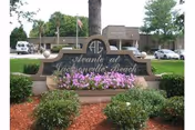 Entrance sign for Avante Villa At Jacksonville Beach with flowers and bushes in front, a tree behind the sign, and a building and parked cars in the background.