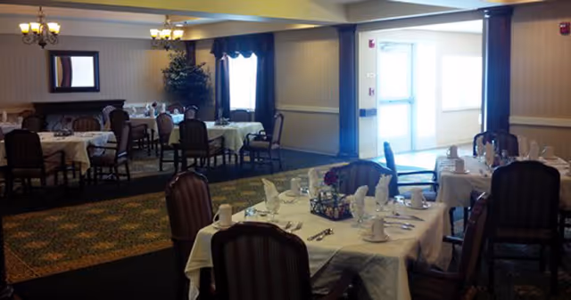 Dining room with multiple tables covered in white tablecloths and set with plates, cups, and napkins, surrounded by upholstered chairs.