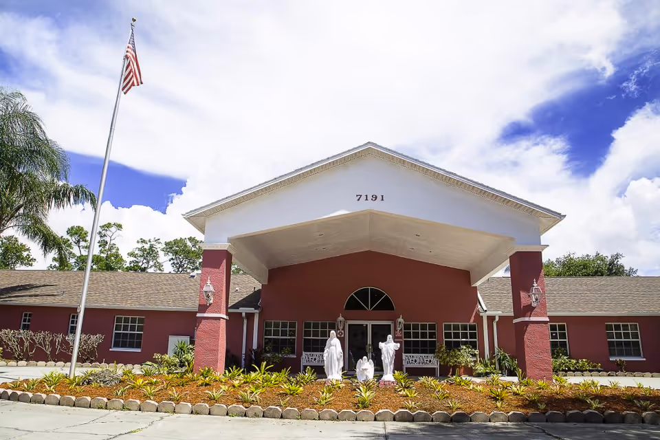 Front view of a single-story assisted living facility with a red exterior, flagpole, and statues.