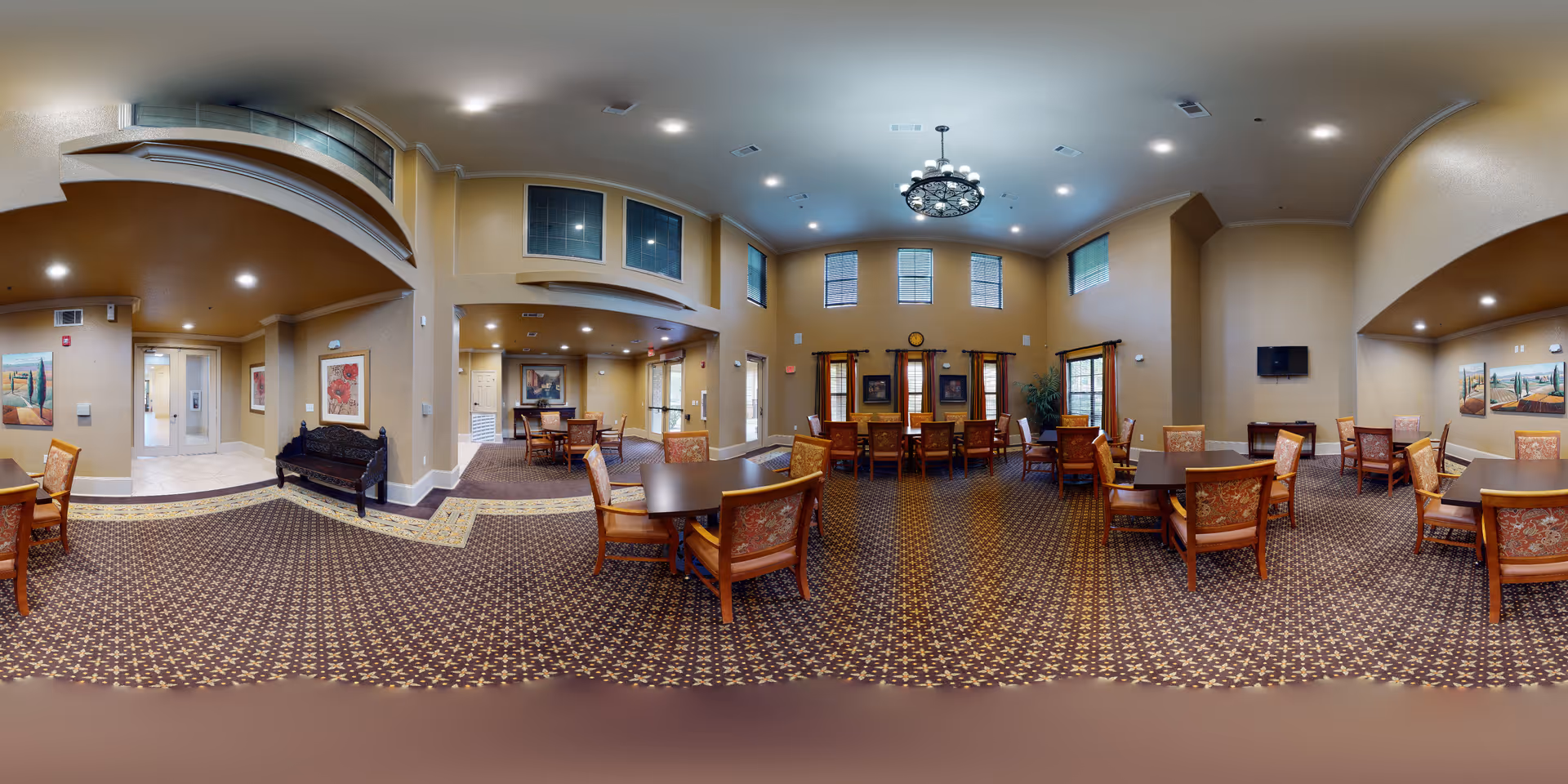 A spacious and well-lit common dining area in a senior living facility with multiple wooden tables and cushioned chairs arranged neatly on a patterned carpet. The room features high ceilings with a decorative chandelier, several windows with curtains, framed artwork on the walls, and a small wall-mounted TV. There is an open hallway leading to other parts of the facility.