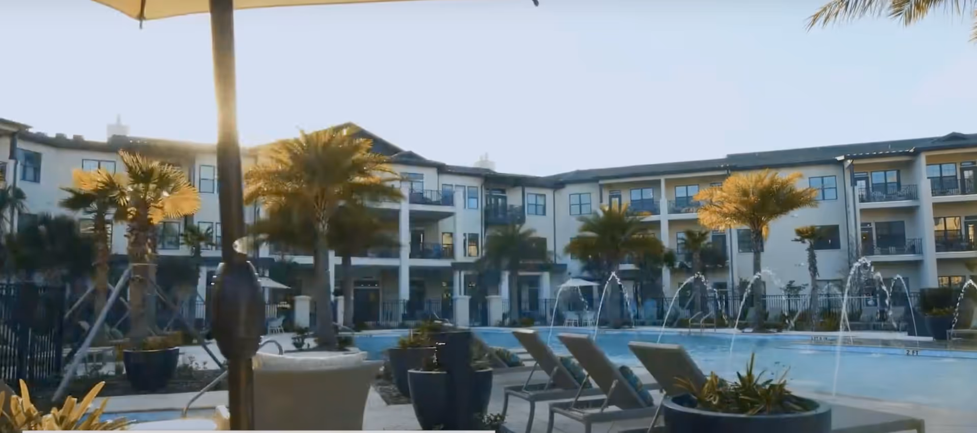 Outdoor courtyard with a swimming pool, lounge chairs, palm trees, fountains, and a multi-story residential building in the background.