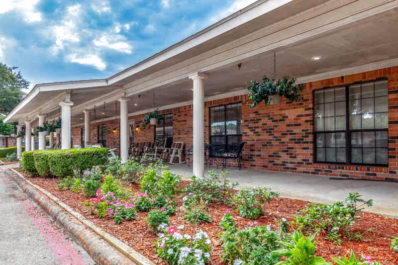 Covered porch area of a brick building with white columns, hanging plants, rocking chairs, and a landscaped flower bed in front under a partly cloudy sky.