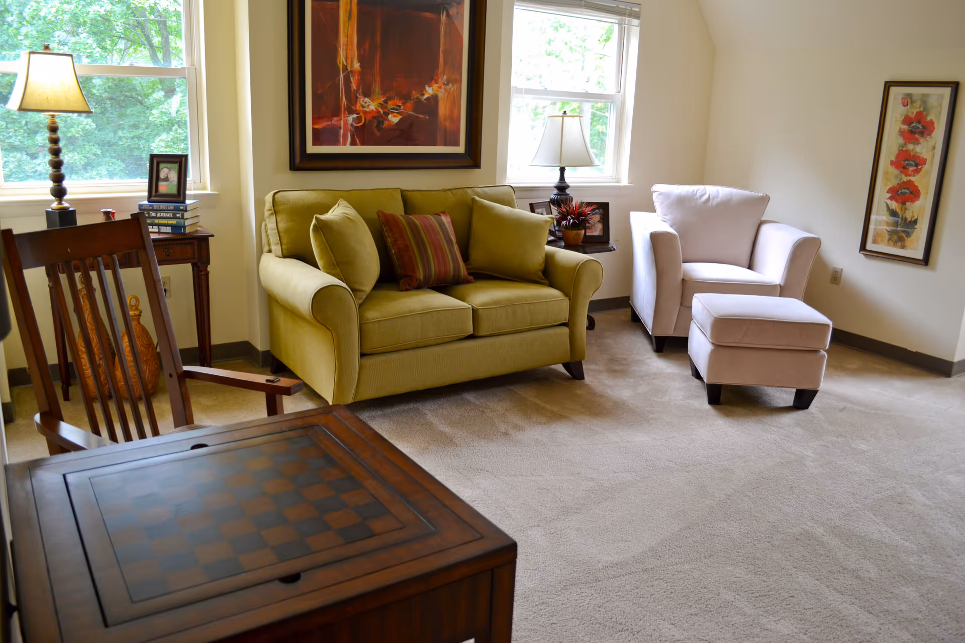 A cozy living room with a green loveseat adorned with three cushions, a beige armchair with a matching ottoman, a wooden chair, and a wooden table with a checkerboard pattern. The room has two windows letting in natural light, a table lamp, framed artwork on the walls, and a small side table with books and a photo frame.