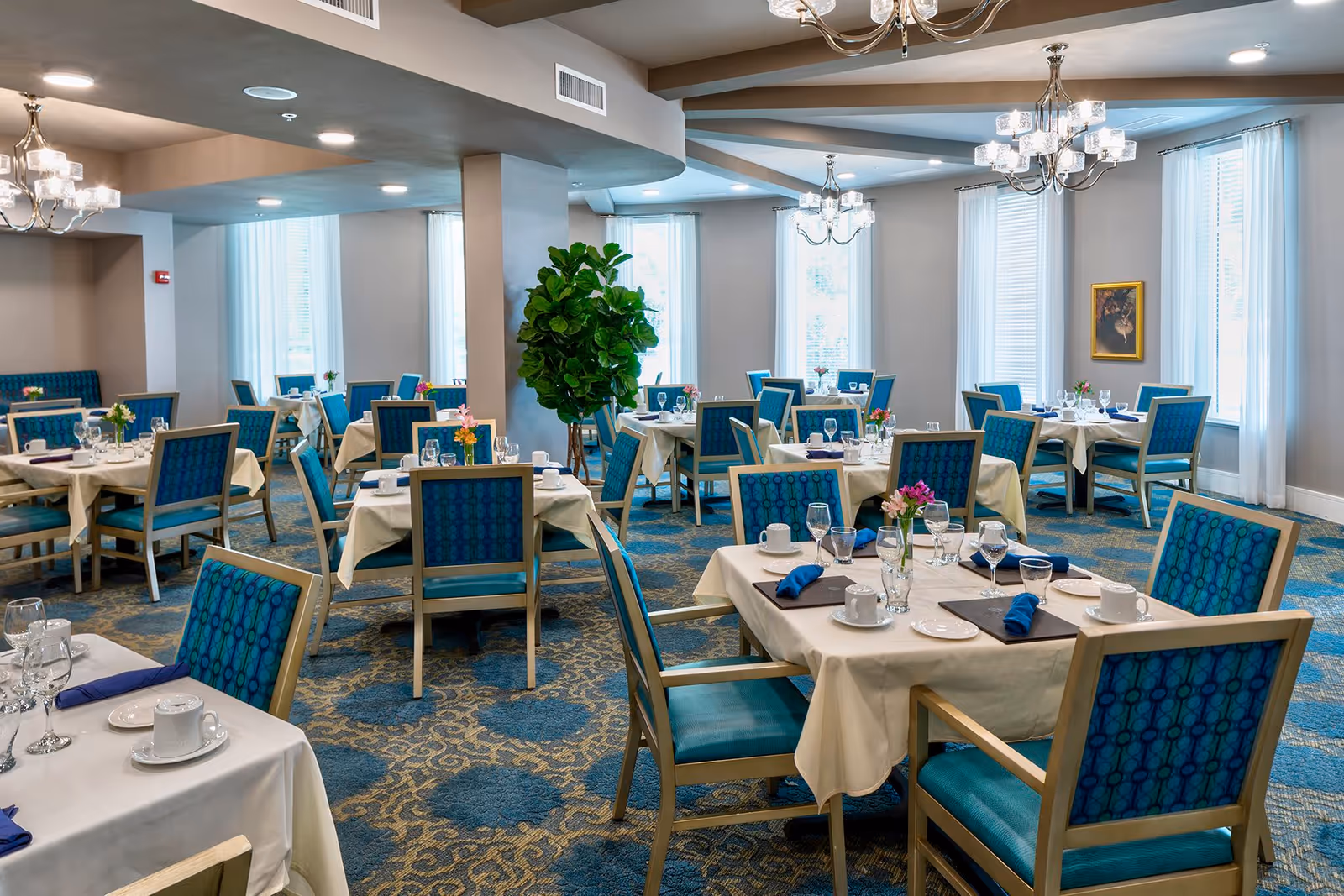 Bright, elegant dining room with tables set with white tablecloths, blue upholstered chairs, chandeliers, and large windows.