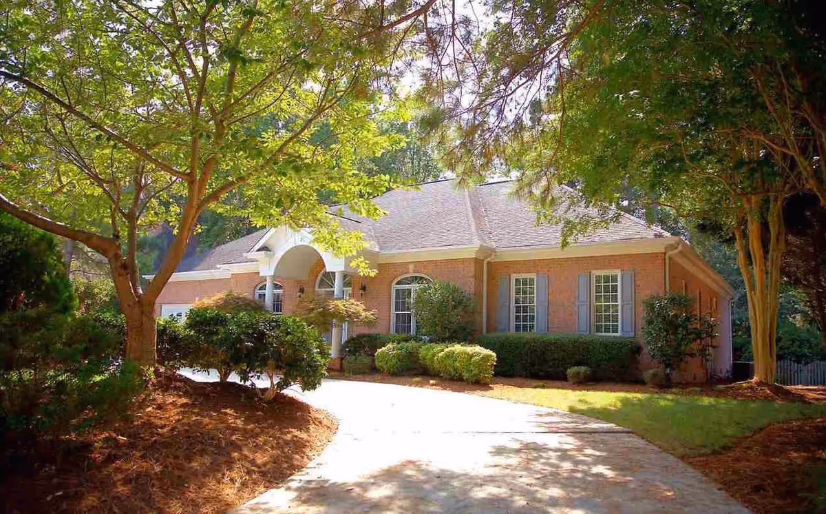 A single-story brick building with a light-colored roof, surrounded by well-maintained shrubs and trees. A curved driveway leads up to the entrance, which features an arched portico with white columns. The scene is bright and sunny with green foliage providing shade.