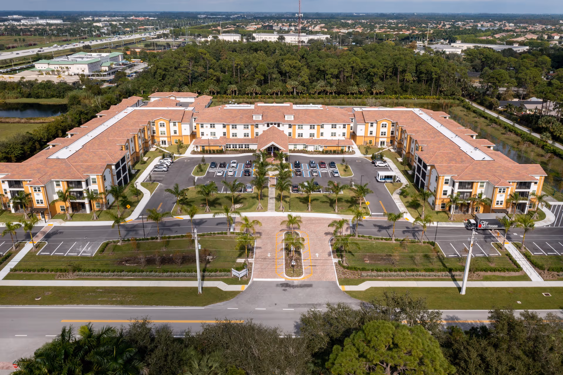 Aerial view of a large U-shaped three-story retirement community building with a central entrance, parking lot, and palm-lined driveway.