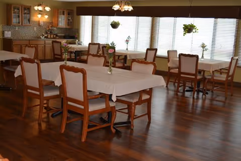 A dining room with wooden floors and several tables covered with white tablecloths. Each table is surrounded by wooden chairs with cushioned seats. The room has large windows with blinds, and hanging plants are suspended near the windows. In the background, there is a counter with cabinets and a tiled backsplash.