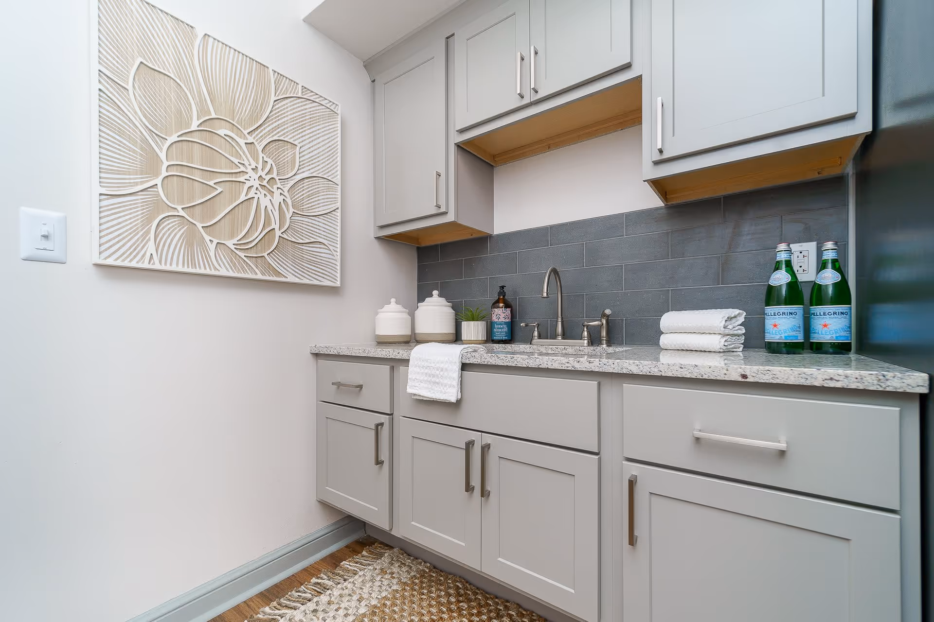 A small kitchen area with light gray cabinets, a granite countertop, and a stainless steel sink with a faucet. On the countertop are two white canisters, a soap dispenser, a small plant, folded white towels, and three bottles of San Pellegrino sparkling water. The backsplash is made of dark gray subway tiles. A decorative wall art piece featuring a large flower design is mounted on the adjacent wall. A woven rug is on the wooden floor beneath the cabinets.