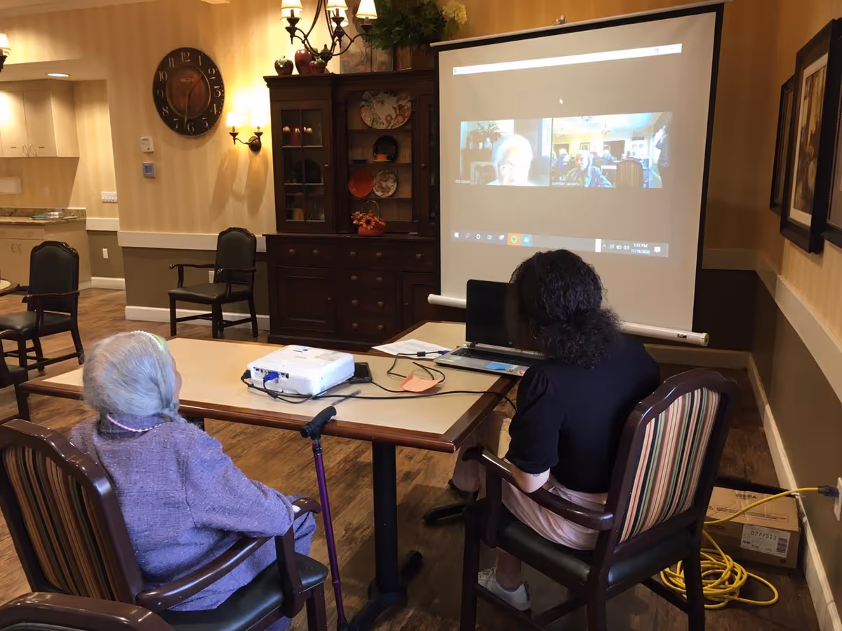 Two women sitting at a table in a cozy room with wooden furniture and warm lighting. One woman has gray hair and is wearing a purple sweater, while the other has dark curly hair and is wearing a black top and light-colored skirt. They are looking at a projection screen displaying a video call with multiple participants. A projector and a laptop are on the table, and a walking cane rests against one chair.