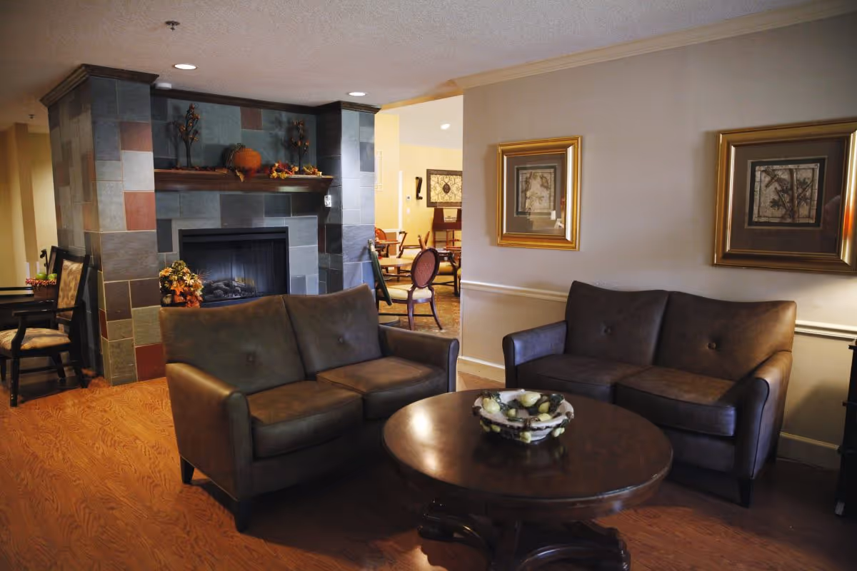 A cozy living room area with two brown leather sofas facing a round wooden coffee table. Behind the sofas is a fireplace with a tiled surround decorated with autumn-themed items. The walls are adorned with framed artwork, and there are additional chairs and tables visible in an adjoining room.