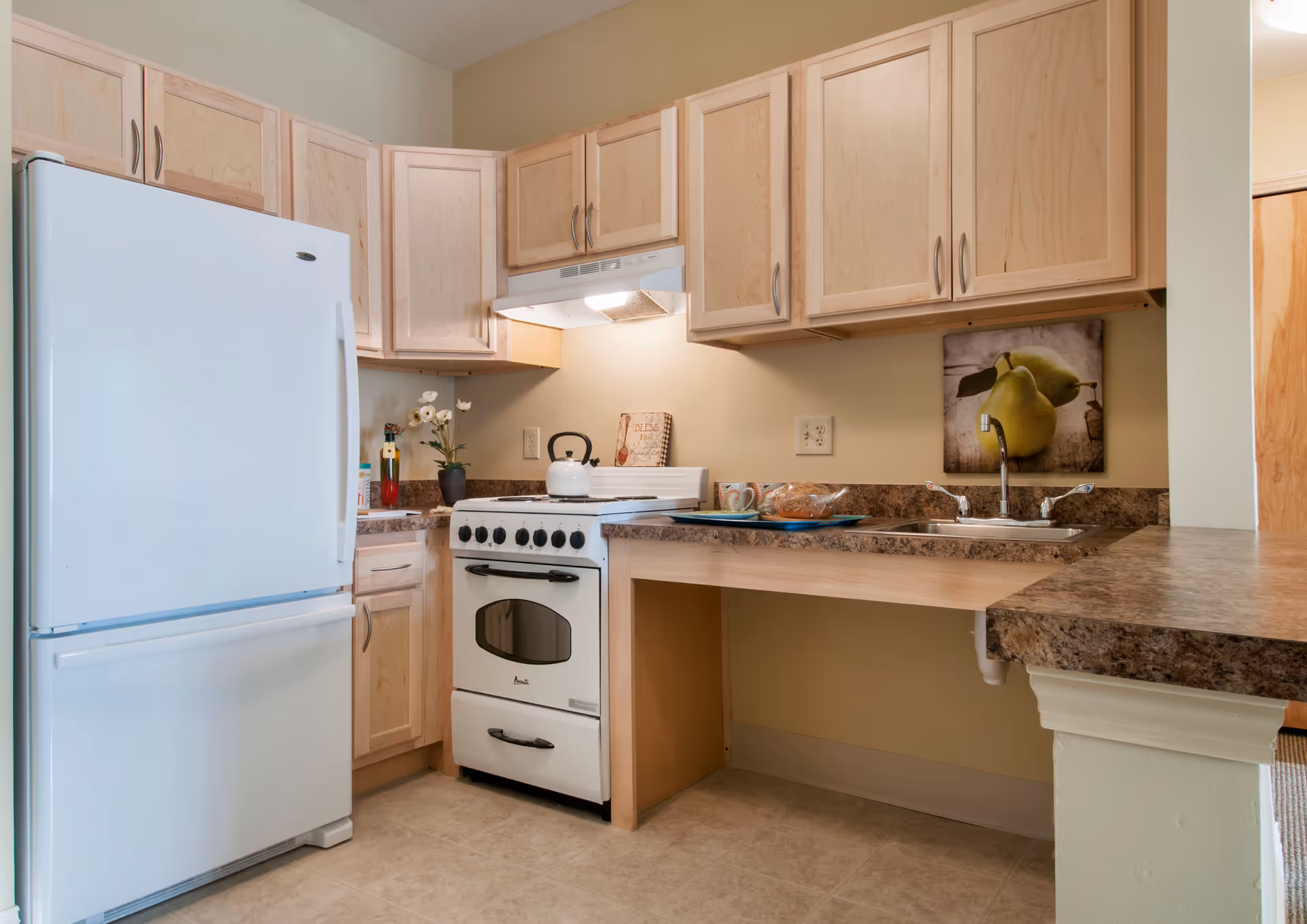 A compact kitchen with light wood cabinets, a white refrigerator, a white stove with a kettle on top, a double sink, and a countertop with a loaf of bread and two mugs. There is a small decorative plant and a wall art featuring pears above the sink.