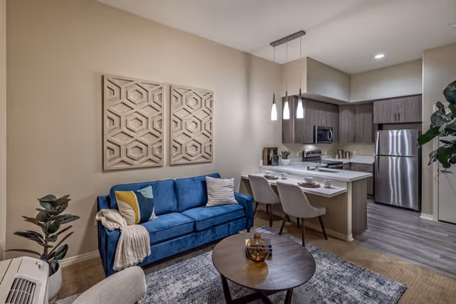 A modern living area featuring a blue sofa with decorative pillows and a throw blanket, a round wooden coffee table with a glass vase, and a kitchen with wooden cabinets, stainless steel appliances, and a breakfast bar with two chairs. The walls are beige with geometric wall art above the sofa, and the floor has a light wood finish.