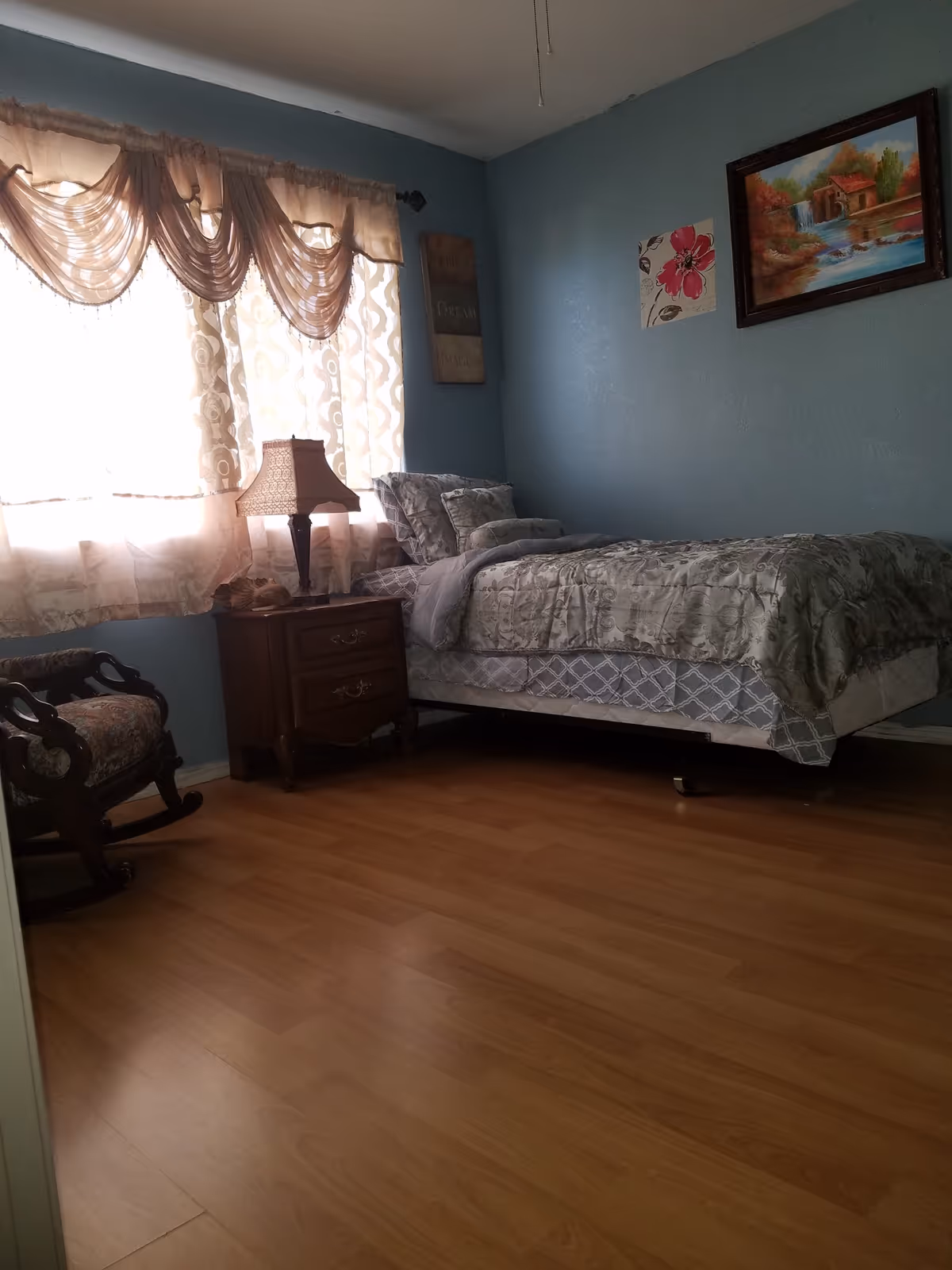 A cozy bedroom with light blue walls, a single bed with patterned bedding, a wooden nightstand with a lamp, a rocking chair with floral upholstery, and a window with sheer curtains letting in natural light. There are two pieces of wall art, one with a floral design and another depicting a landscape with a waterfall.