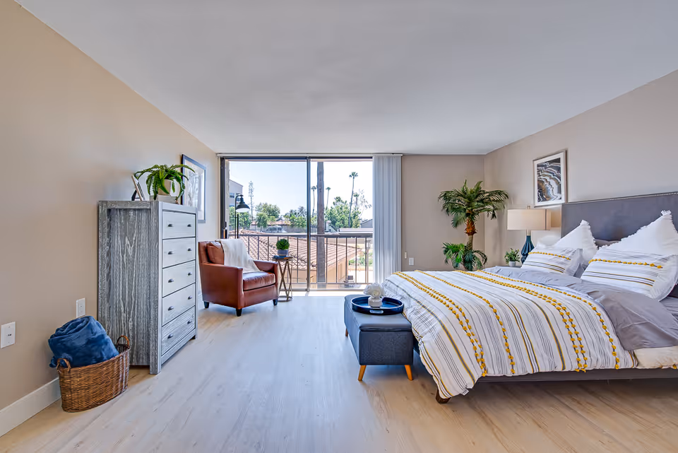 Bright and spacious bedroom with a large bed featuring white and yellow striped bedding, a gray upholstered headboard, a blue ottoman at the foot of the bed, a wooden dresser with a plant on top, a brown leather armchair next to a small side table, and a large sliding glass door leading to a balcony with a view of rooftops and trees outside.