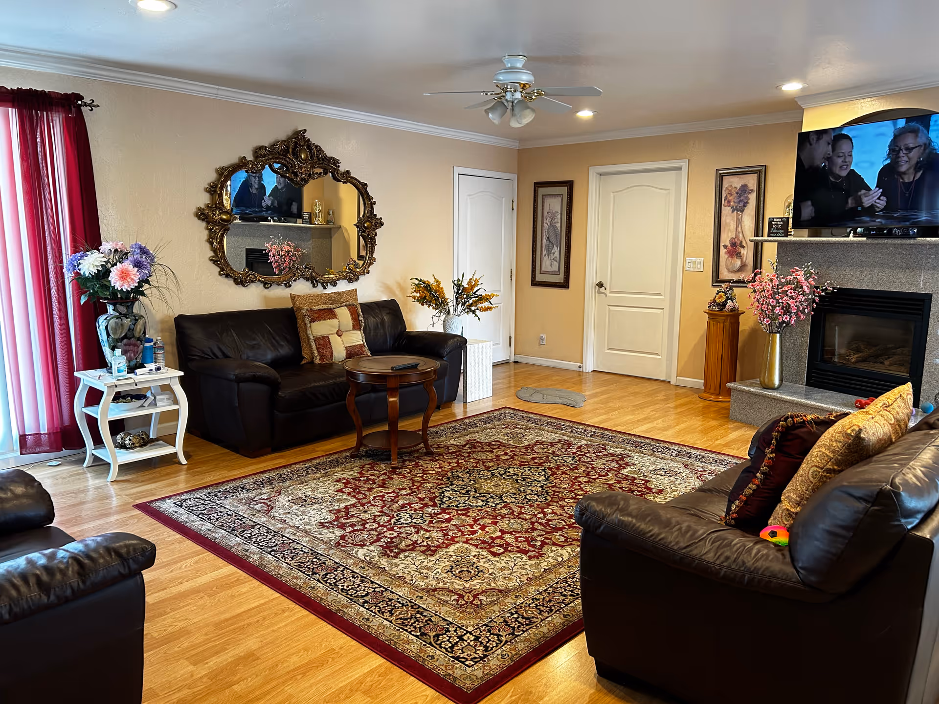 A cozy living room with wooden flooring, two dark leather sofas with decorative pillows, a large ornate mirror on the wall, a patterned area rug in red and beige tones, a small round wooden coffee table, a white side table with a vase of flowers, and a fireplace with a TV mounted above it. The room has beige walls, two closed white doors, framed artwork, and red curtains on the window.