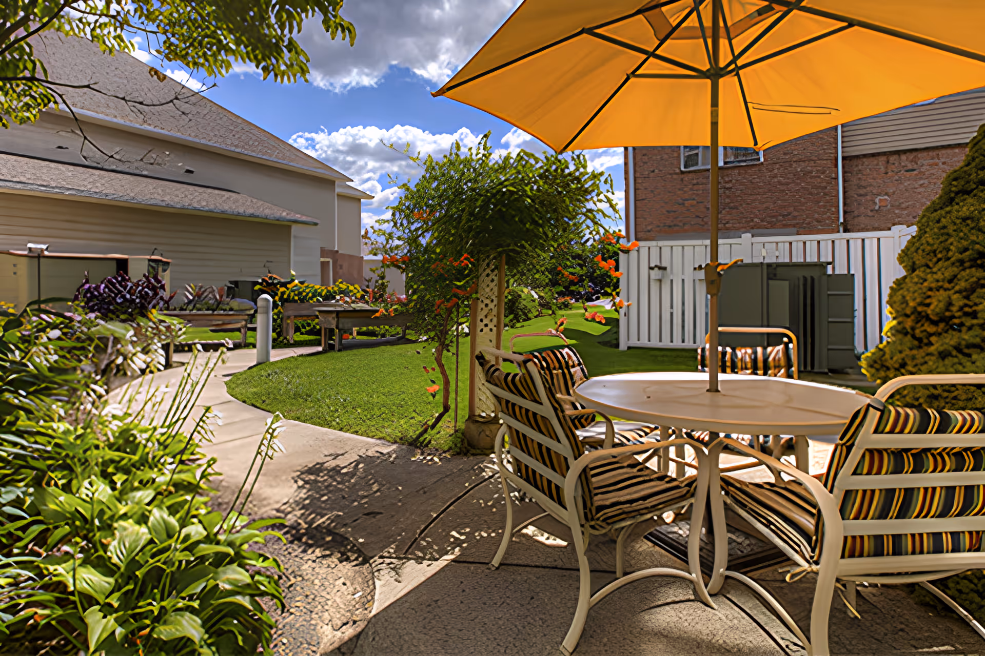 Sunlit outdoor courtyard with a round table and striped chairs under a yellow umbrella, a paved walkway and landscaped greenery.
