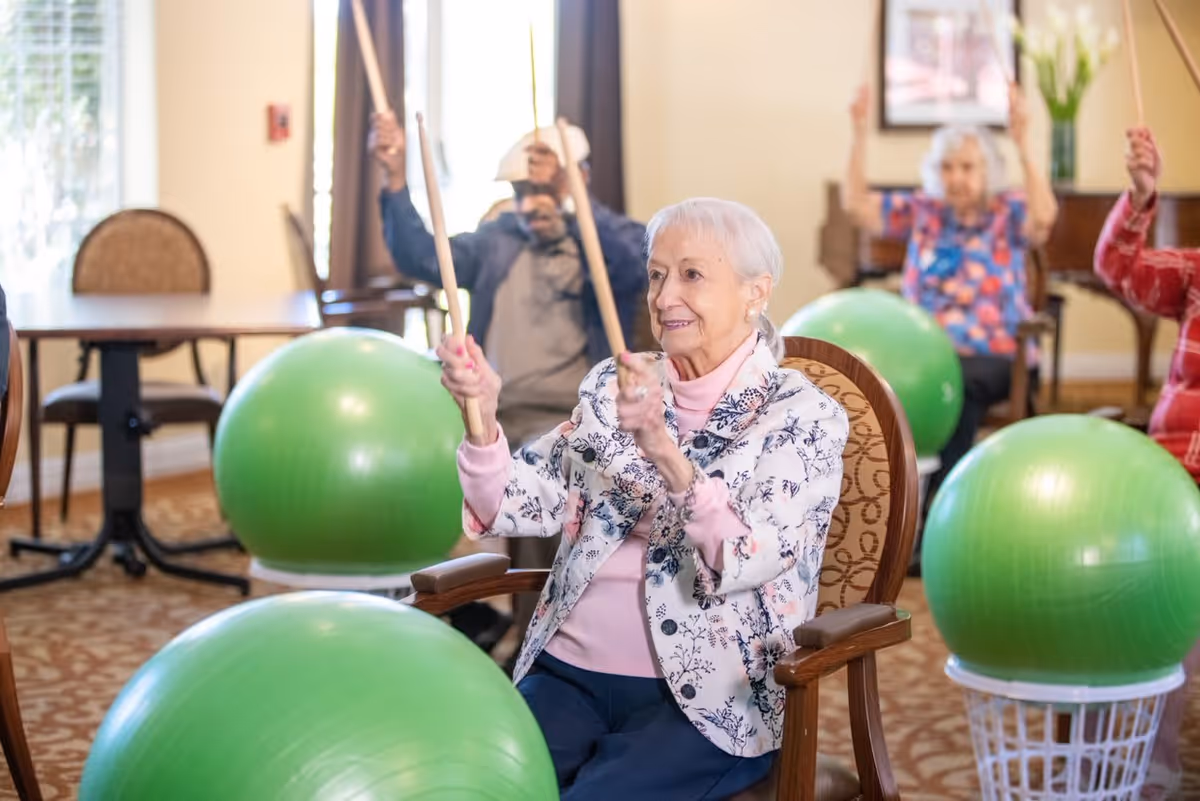 A group of elderly individuals seated in chairs participating in a seated exercise class using green exercise balls and wooden drumsticks in a well-lit room with tables and chairs in the background.