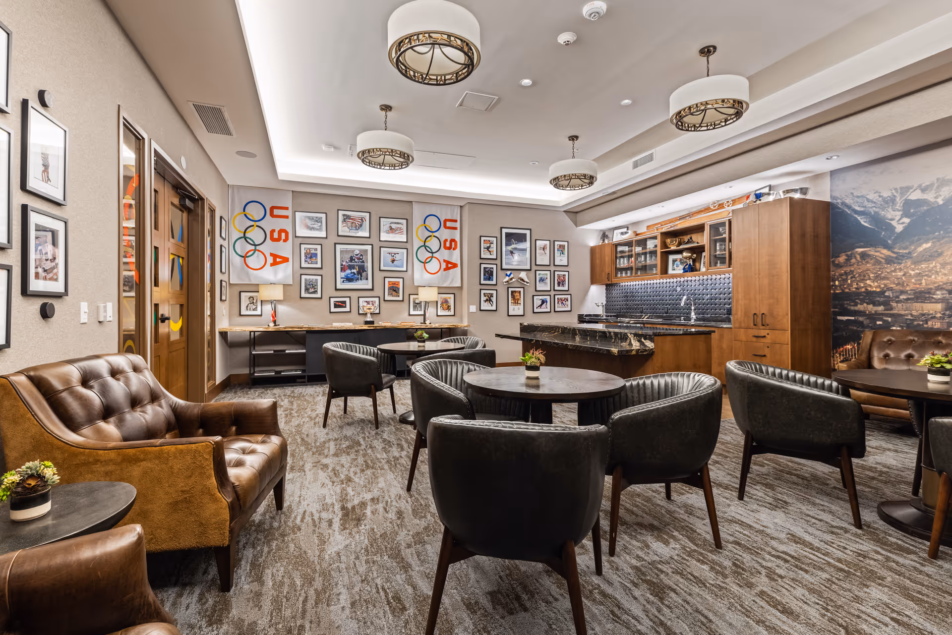 A cozy common area in a senior living facility featuring several round tables with black chairs, a brown leather sofa, and a kitchenette with wooden cabinets and a black countertop. The walls are decorated with framed pictures, including USA Olympic flags and various sports images. The room has modern ceiling lights and a carpeted floor.