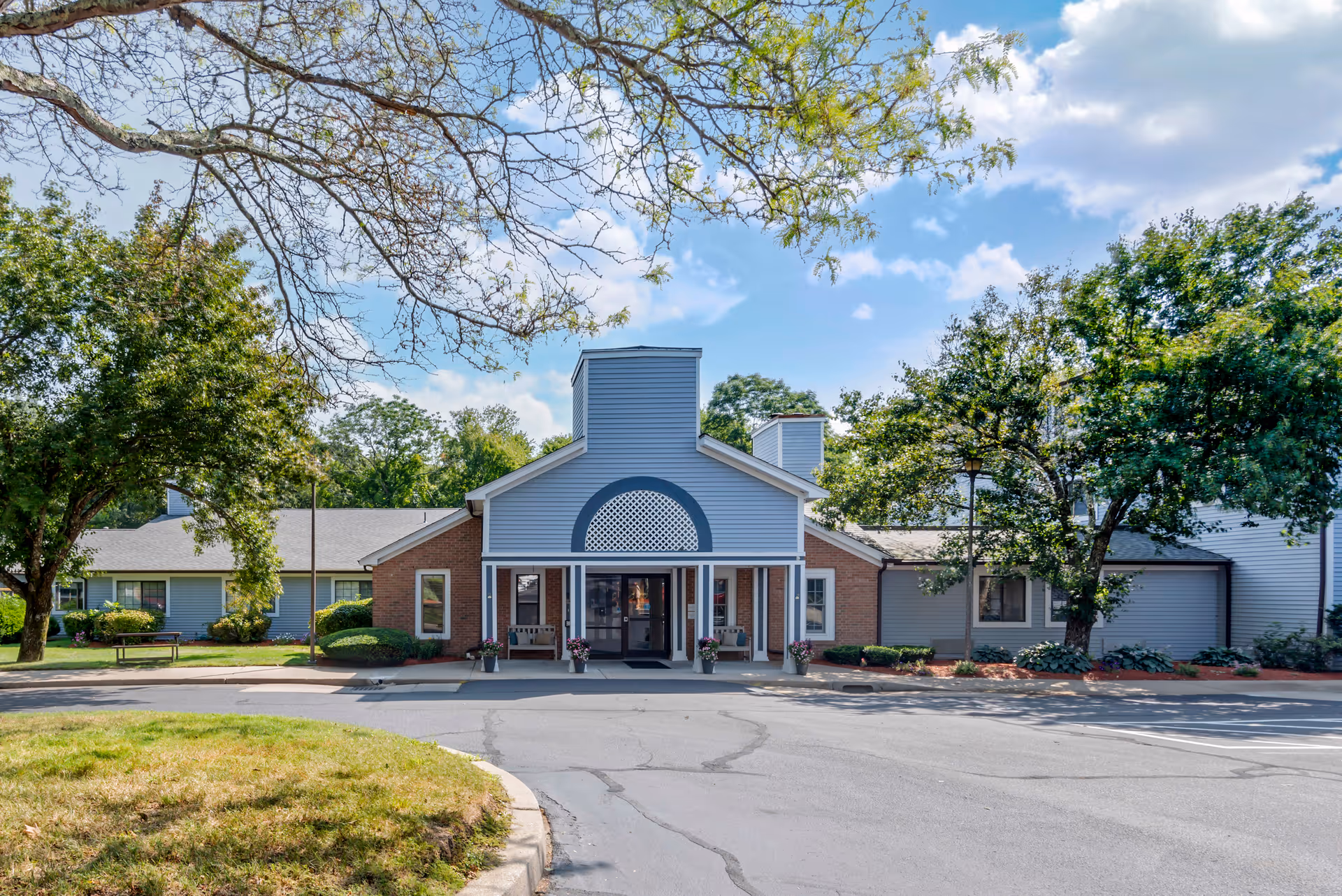 Front exterior view of a single-story senior living facility building with a central entrance featuring a covered porch, surrounded by trees and landscaping under a partly cloudy sky.