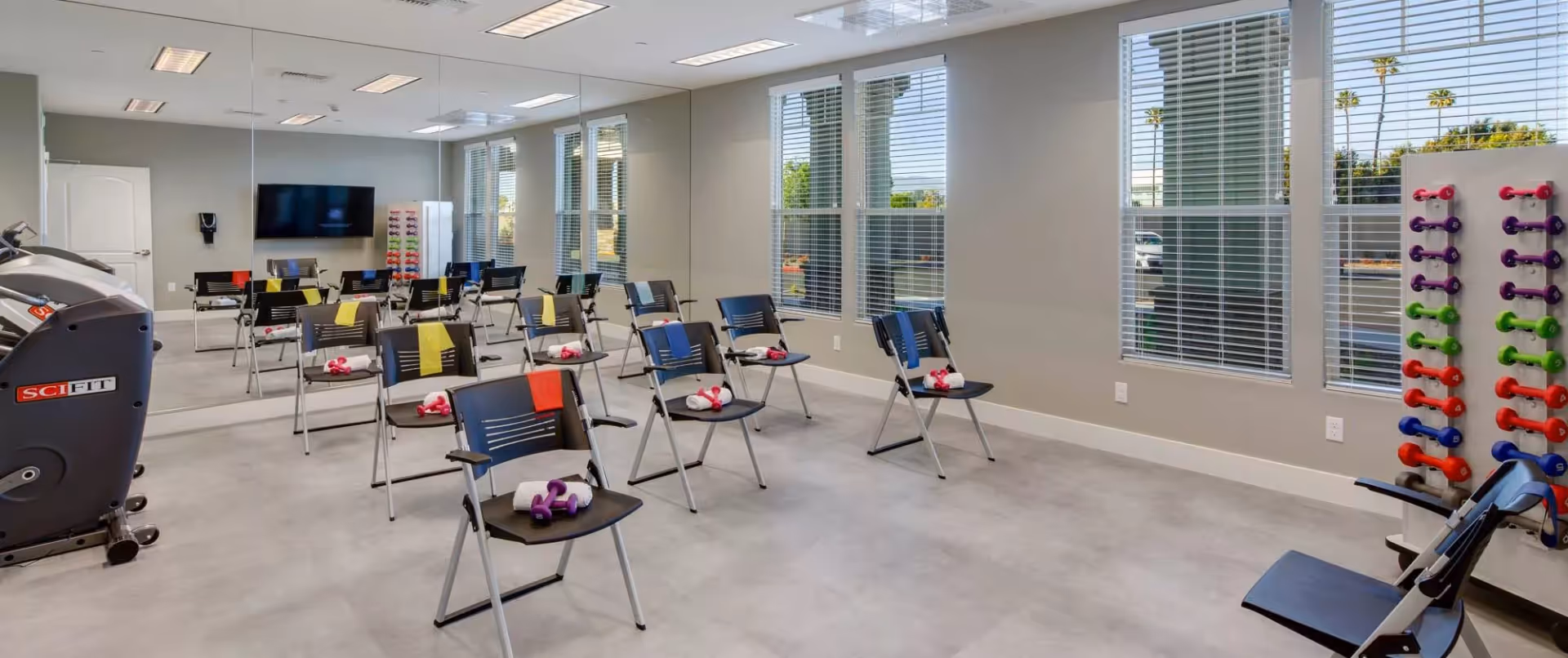 Bright fitness room with rows of chairs, small dumbbells and towels, a mirrored wall, windows, and exercise equipment.
