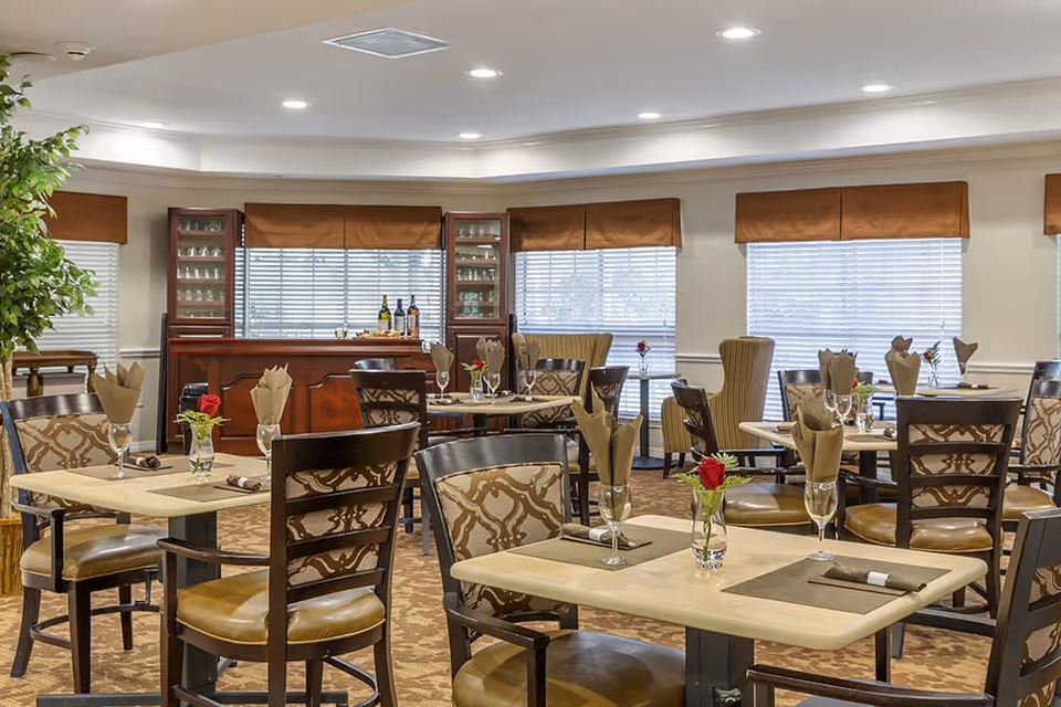 A well-lit dining room with multiple tables set with placemats, napkins, and glasses, each table featuring a small vase with a red rose. The room has patterned chairs, large windows with brown valances, and a wooden cabinet with glass doors displaying glassware and bottles.