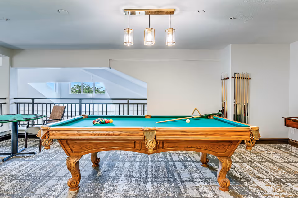 A well-lit game room featuring a wooden pool table with green felt, pool balls arranged in a triangle, and a cue stick resting on the table. The room has a patterned carpet, a modern three-light ceiling fixture, a small table with two chairs to the left, and a rack holding multiple pool cues on the right wall. A railing and windows with natural light are visible in the background.