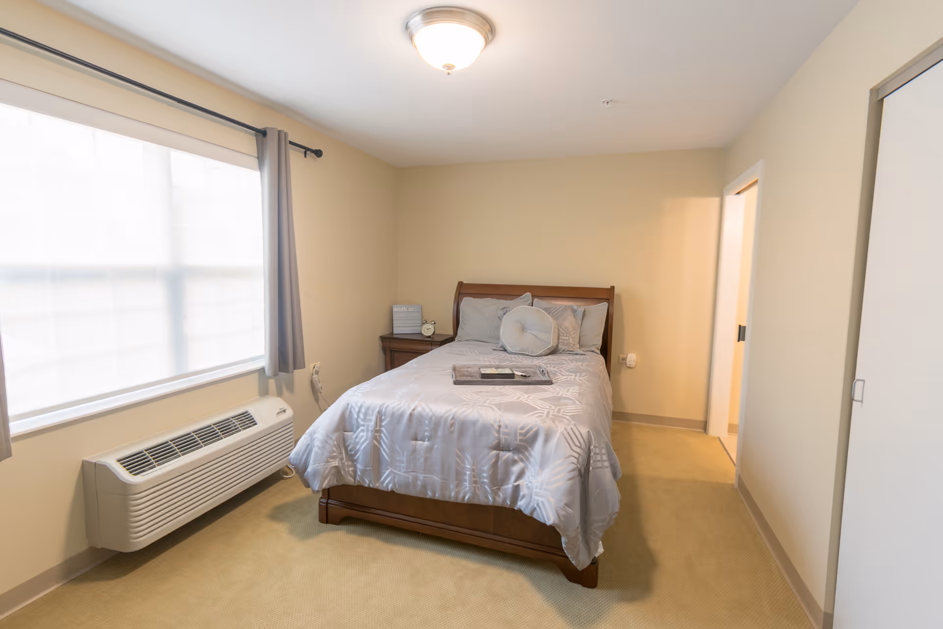 A small, neatly arranged bedroom with a double bed covered in a light gray patterned comforter and multiple pillows. There is a wooden nightstand with a clock and a small decorative item next to the bed. A large window with sheer white curtains and gray drapes lets in natural light. The room has beige walls and carpet, a ceiling light fixture, and a white air conditioning unit below the window. A doorway leads to another room or hallway.