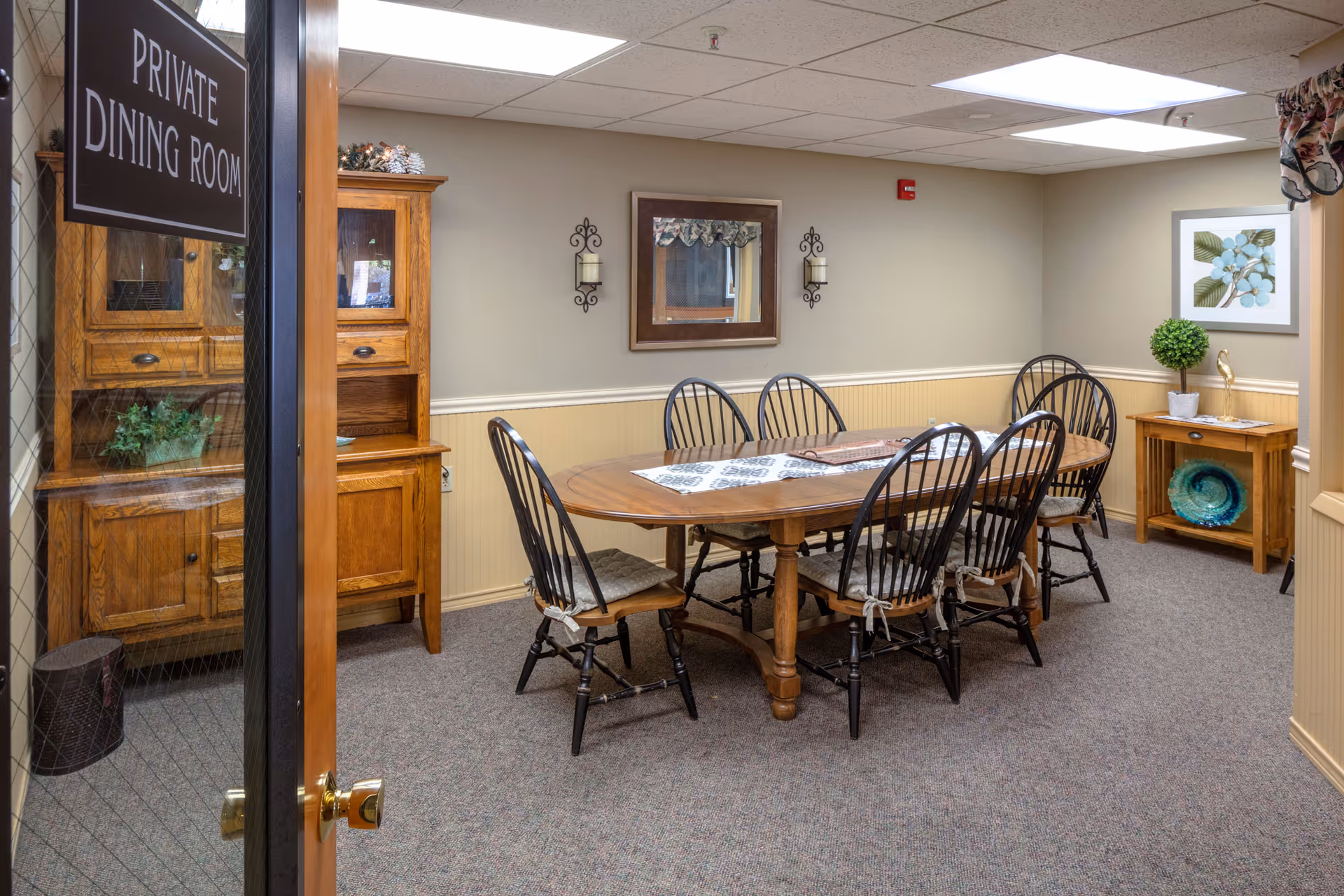Interior view of a private dining room with a wooden dining table surrounded by six black chairs with cushions. The room has beige walls with a chair rail, a wooden cabinet with glass doors, a small wooden side table with a plant and decorative plate, a framed floral picture on the wall, and two wall-mounted candle holders flanking a mirror. The door to the room has a sign that reads 'PRIVATE DINING ROOM'.