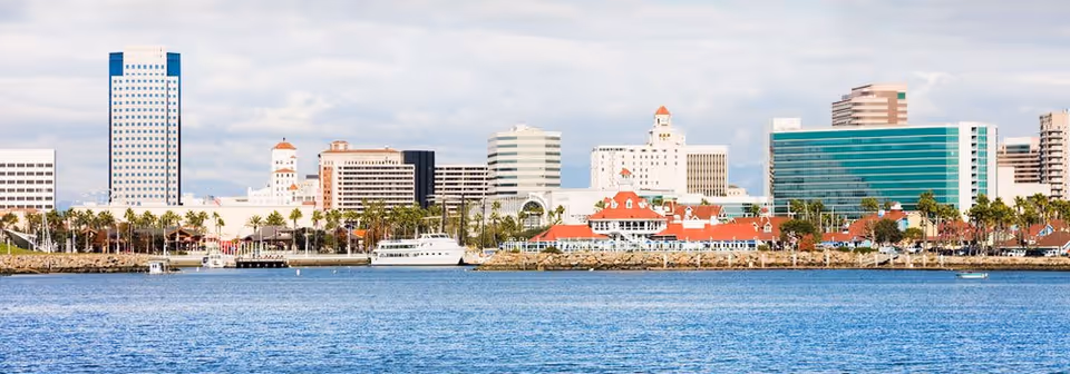 Panoramic view of the Long Beach waterfront skyline with various buildings, palm trees, and a large body of water in the foreground under a partly cloudy sky.