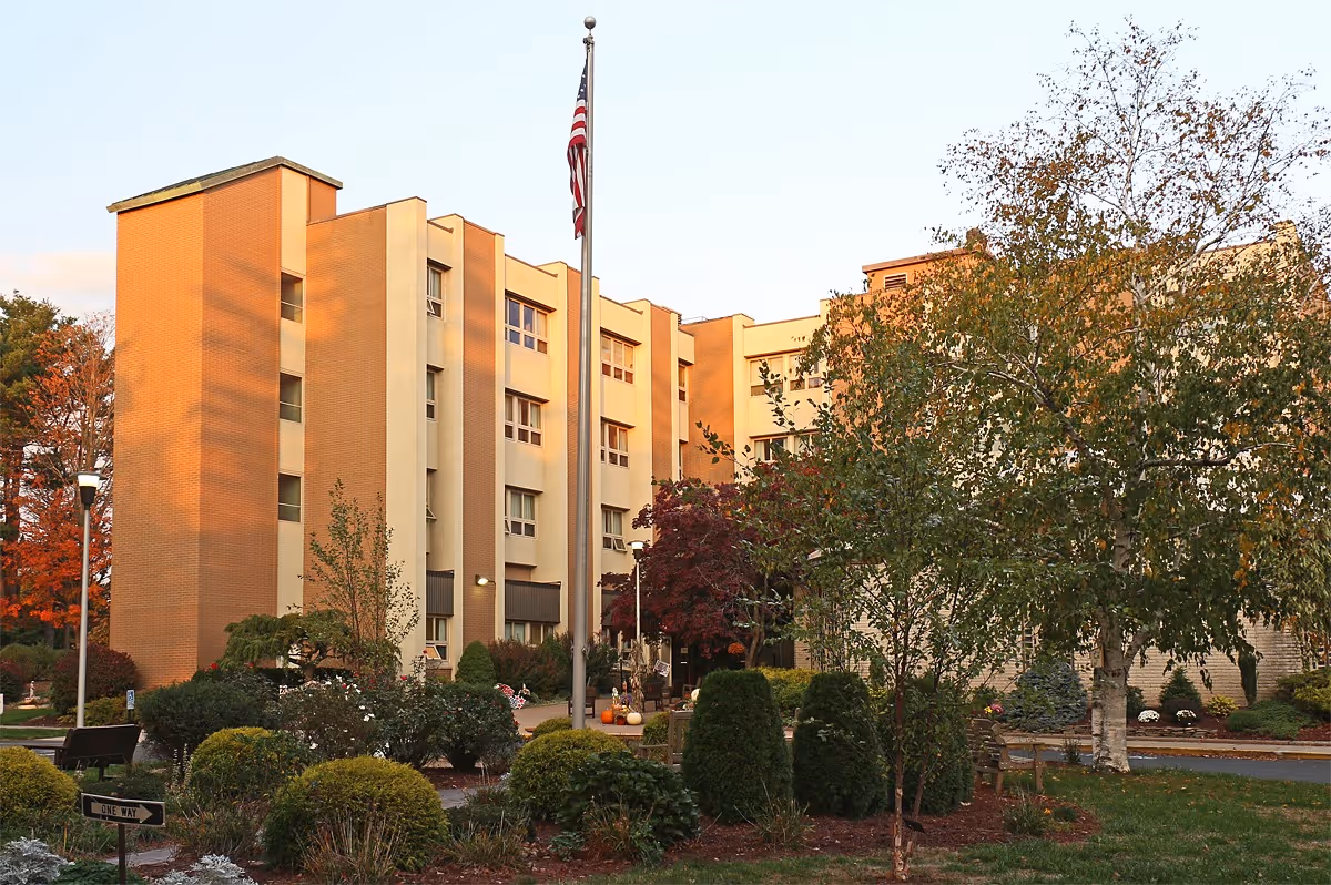 Exterior view of a multi-story senior living facility building with beige and light brown walls, surrounded by landscaped bushes, trees, and a flagpole with an American flag in front.