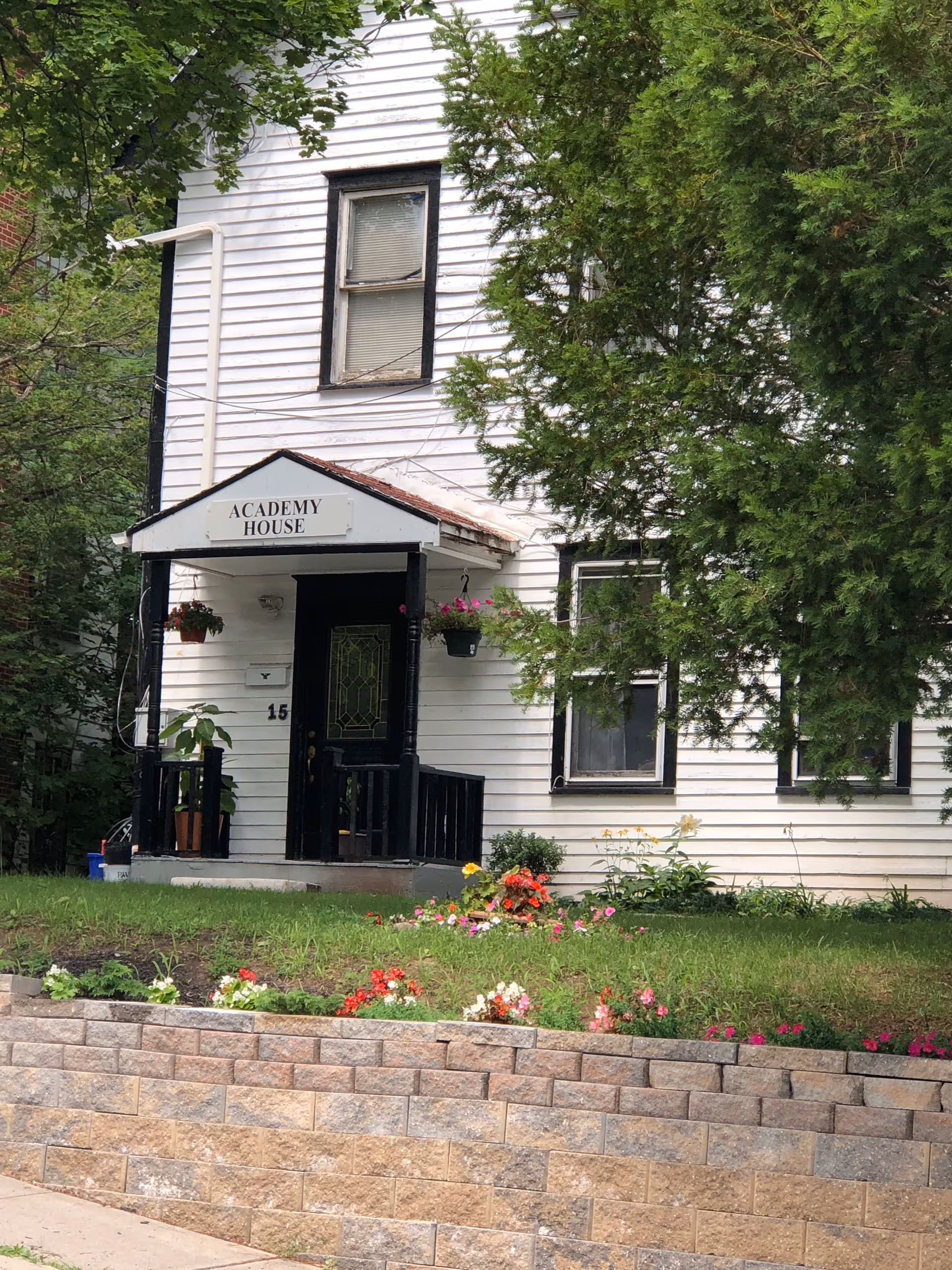 Front exterior view of a white two-story house with black trim named Academy House. The house has a small porch with black railings, hanging flower pots, and a garden with colorful flowers in front. There are trees partially obscuring the right side of the house.